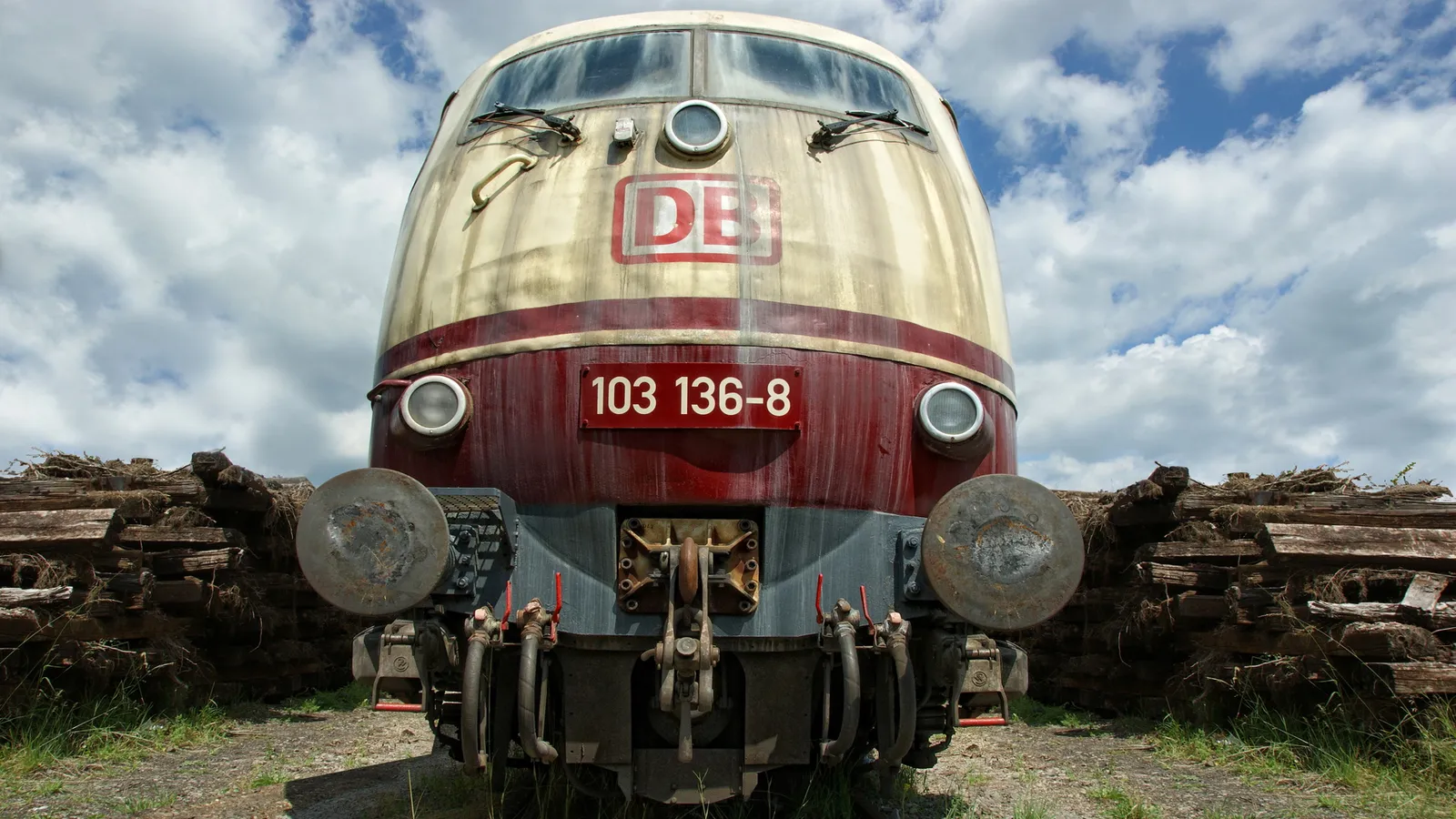 The front of a weathered red and cream DB train engine sits idle, surrounded by piles of old wooden beams.