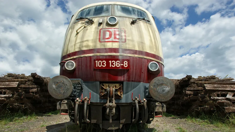 The front of a weathered red and cream DB train engine sits idle, surrounded by piles of old wooden beams.