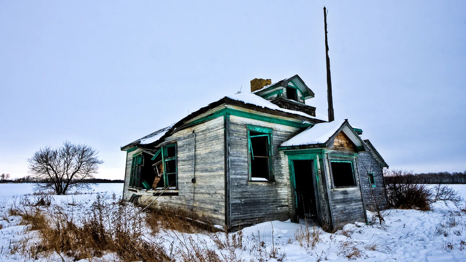 An old, weathered wooden house with broken windows sits in a snowy field under a pale sky.