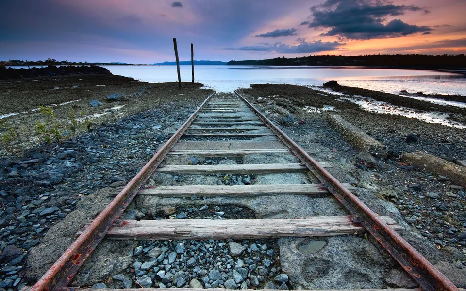 Old railway tracks leading towards a calm sea under a colorful sunset sky.