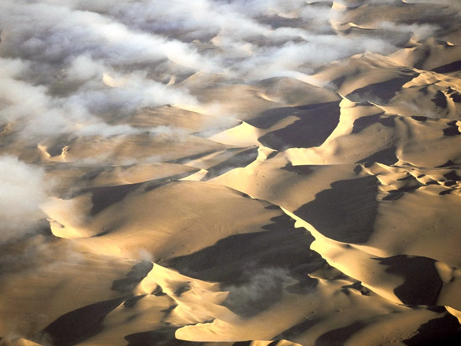 An aerial view of vast sand dunes with shadows and patches of clouds drifting over them.
