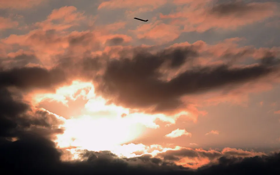 A silhouette of an airplane flies across a sky filled with dramatic, colorful sunset clouds.