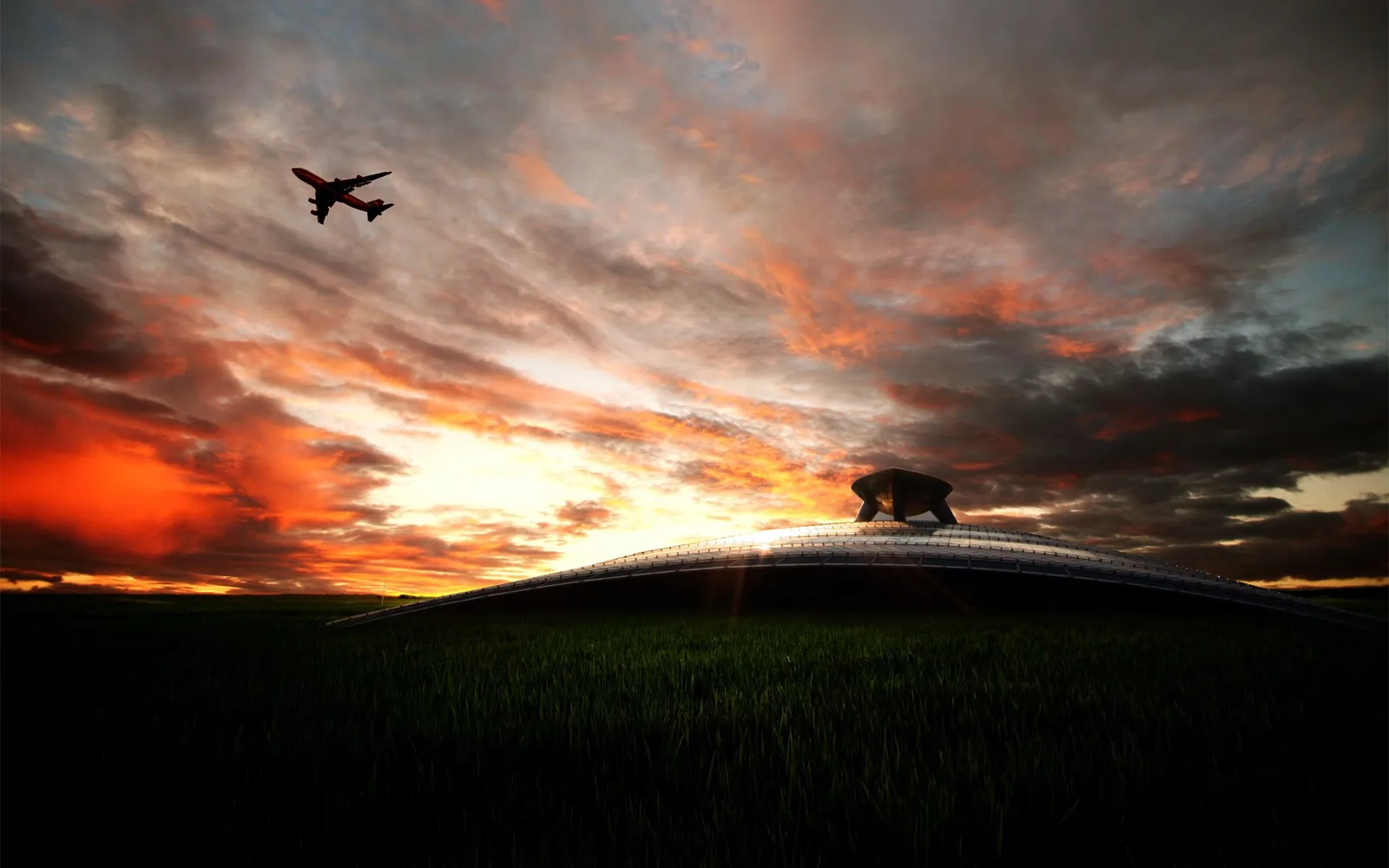 An airplane flies across a vibrant orange and red sunset sky over a grassy field and a futuristic building.