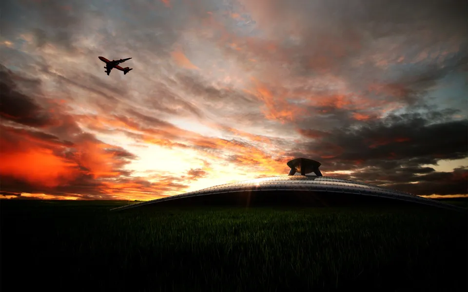 An airplane flies across a vibrant orange and red sunset sky over a grassy field and a futuristic building.