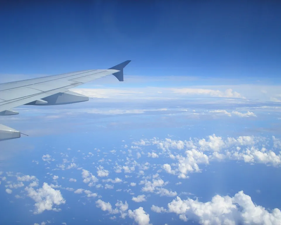View from an airplane window showing the wing and a vast expanse of blue sky with scattered white clouds.