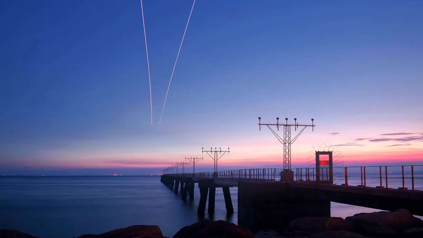 A pier with airport approach lights stretches into the ocean under a colorful twilight sky with contrails.
