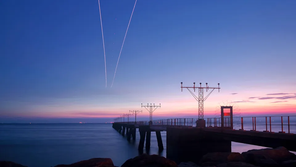 A pier with airport approach lights stretches into the ocean under a colorful twilight sky with contrails.
