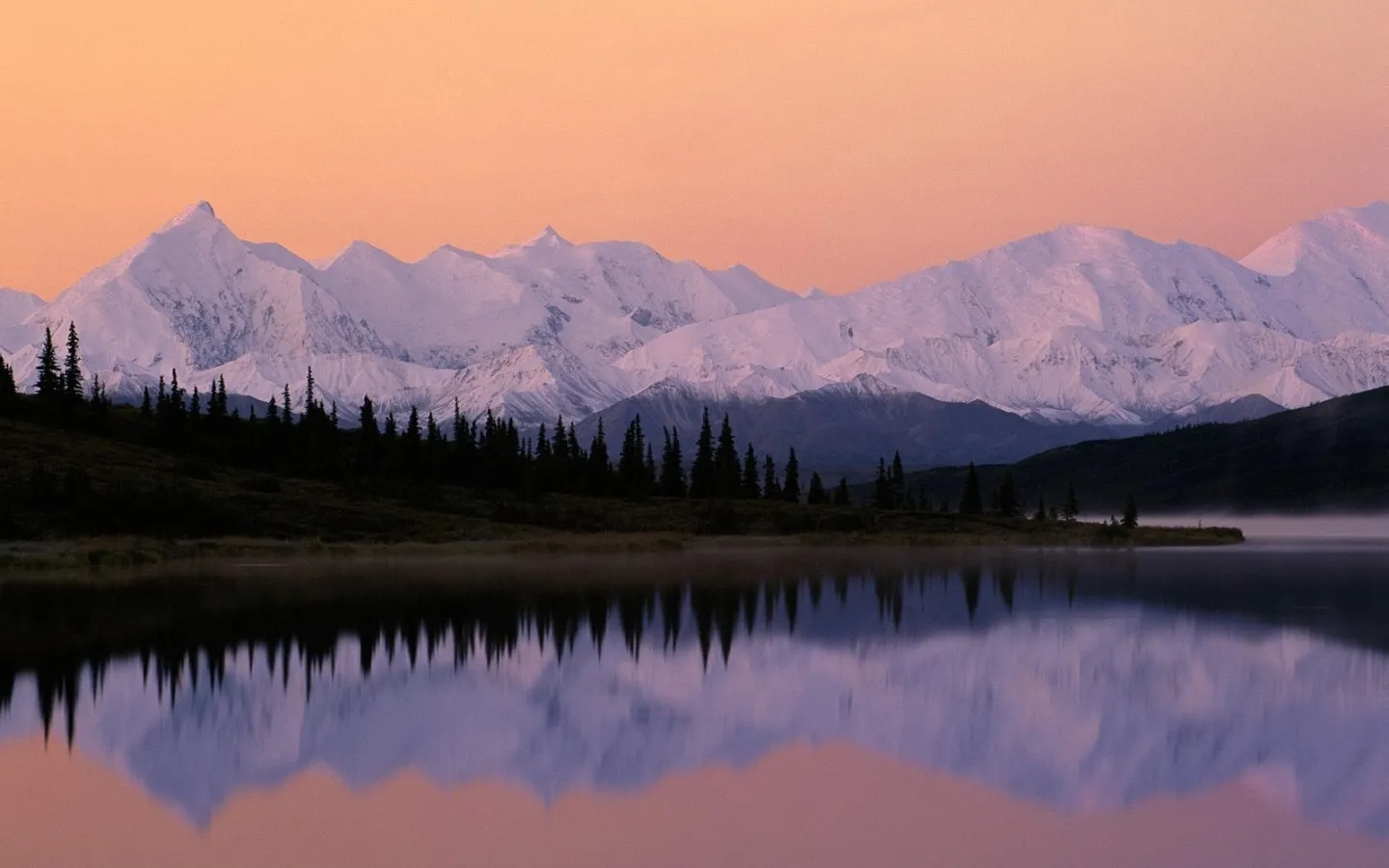 Snowy mountains reflect in a misty lake at sunrise.