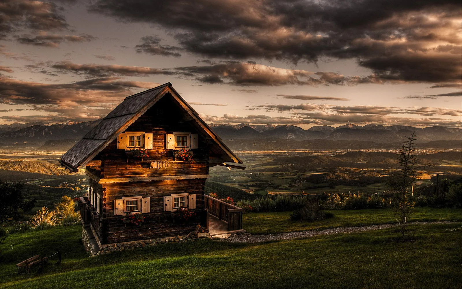 A wooden cabin sits on a grassy hill with mountains in the background during a dramatic sunset.