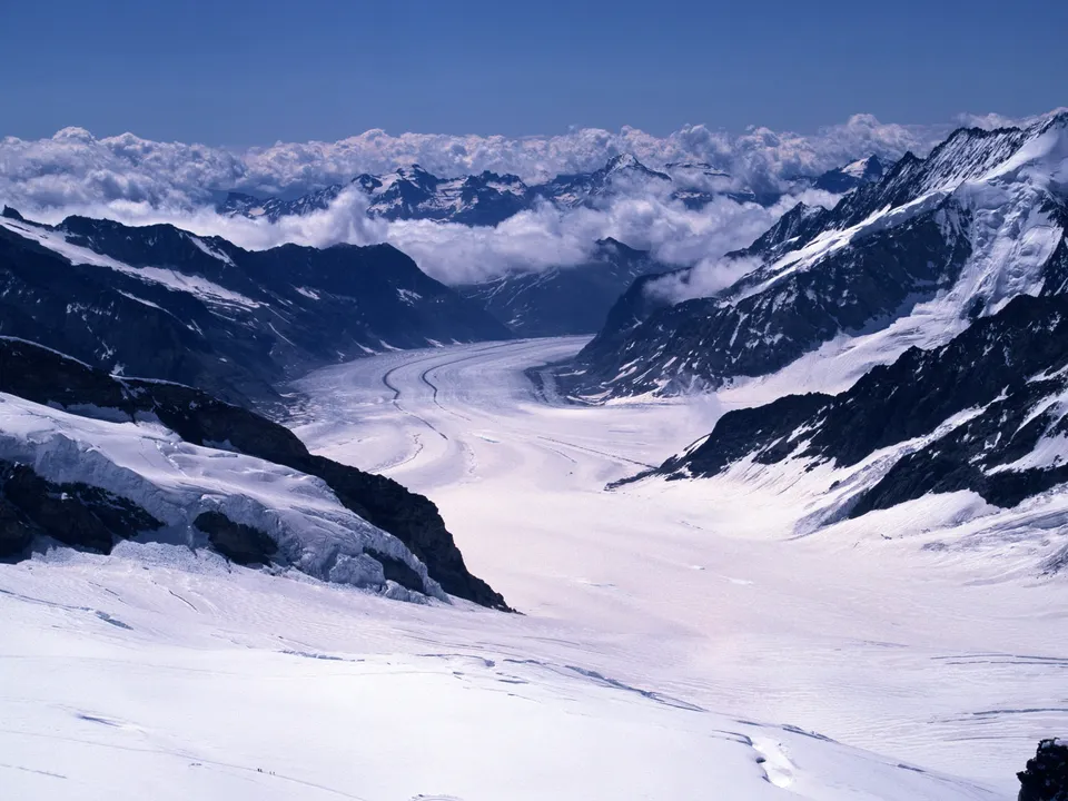 A vast, snow-covered glacier valley winds between towering, snow-capped mountains under a bright blue sky with scattered clouds.