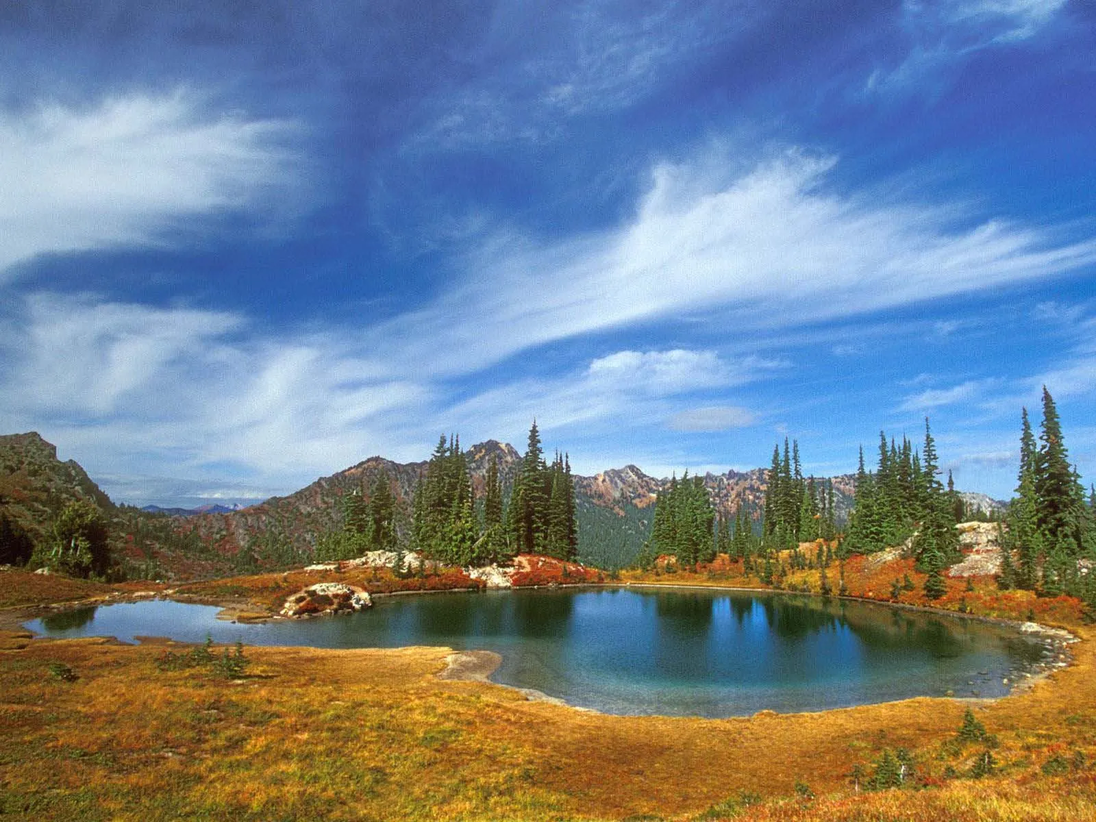 A calm alpine lake reflects the blue sky and surrounding evergreen trees and autumn-colored vegetation.