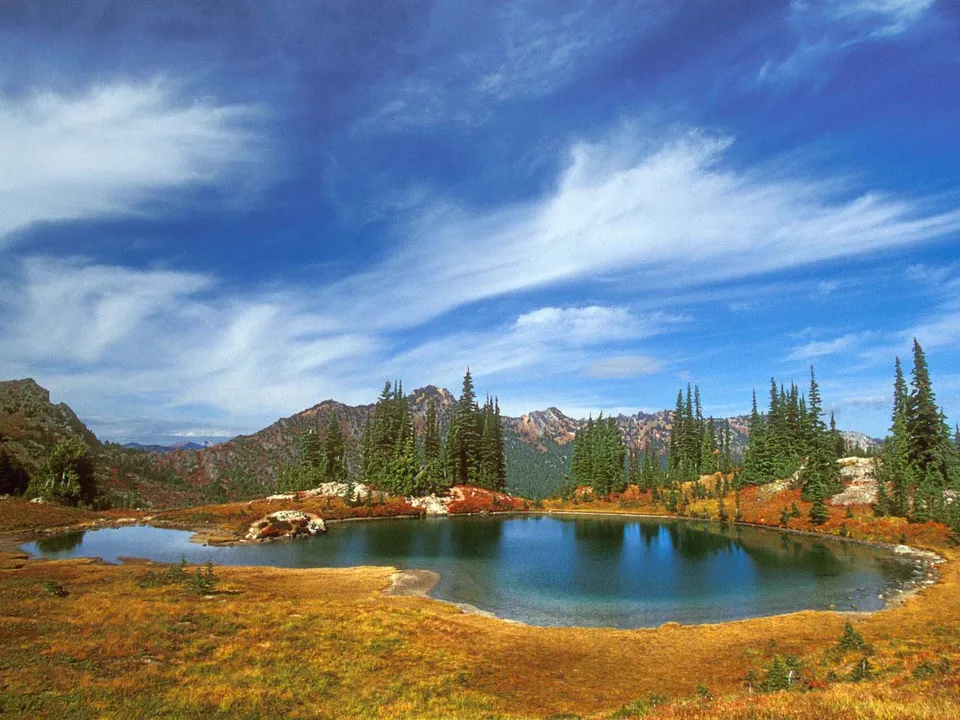 A calm alpine lake reflects the blue sky and surrounding evergreen trees and autumn-colored vegetation.