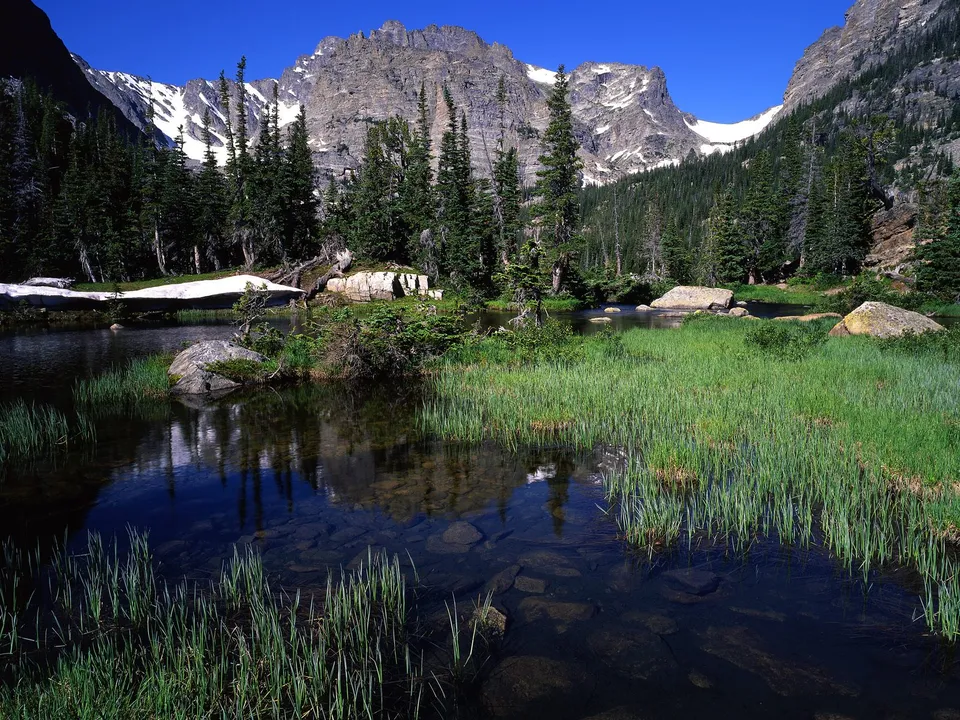 A clear alpine lake reflects snow-capped mountains and evergreen trees under a bright blue sky.