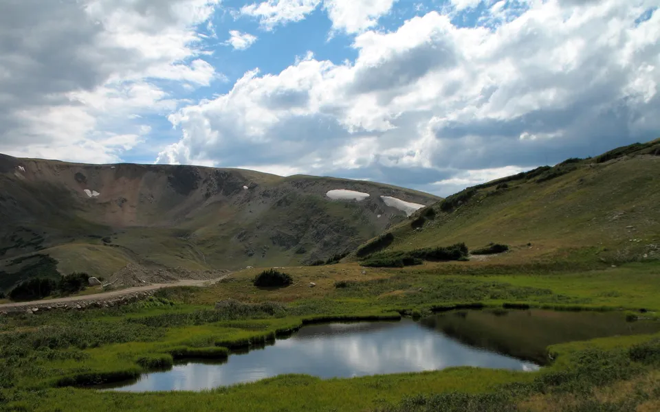 A small alpine lake in the foreground reflects the sky and surrounding green hills and mountains under a cloudy sky.