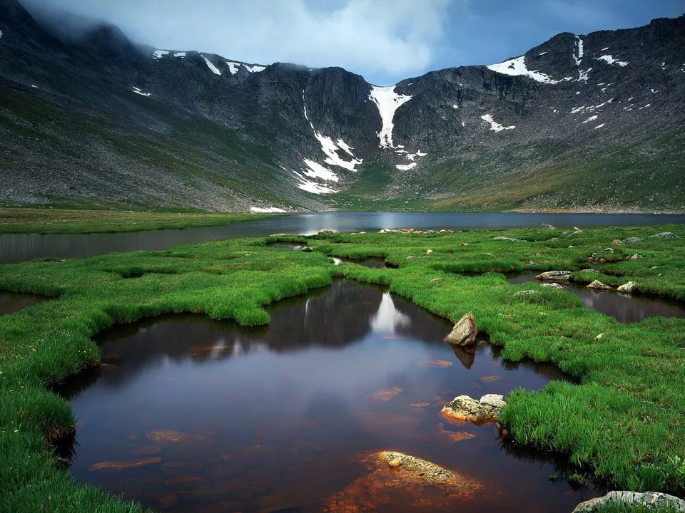 A calm alpine lake reflects the surrounding mountains and green grassy banks under a cloudy sky.