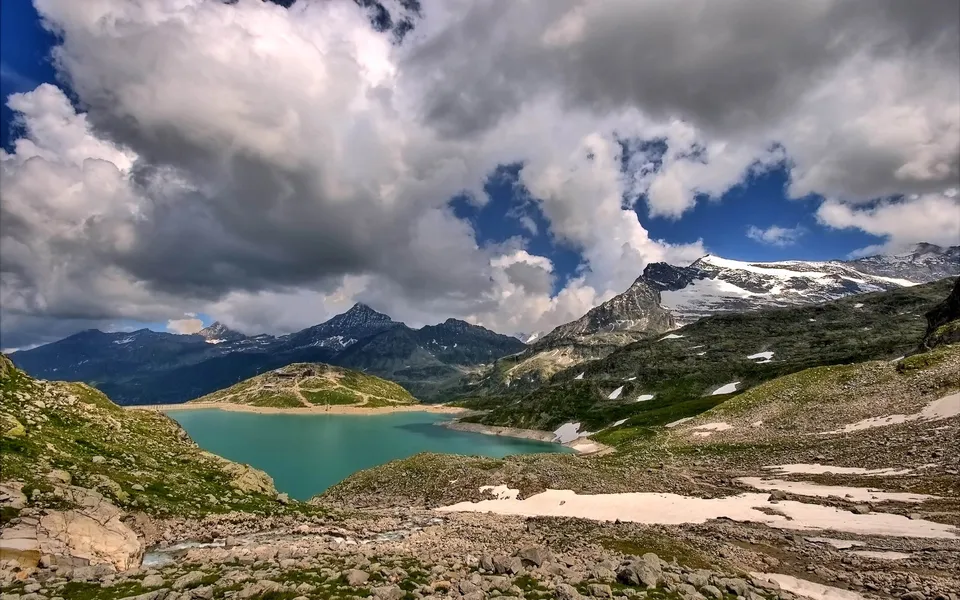A vibrant turquoise alpine lake is nestled among rugged mountains under a dramatic cloudy sky.
