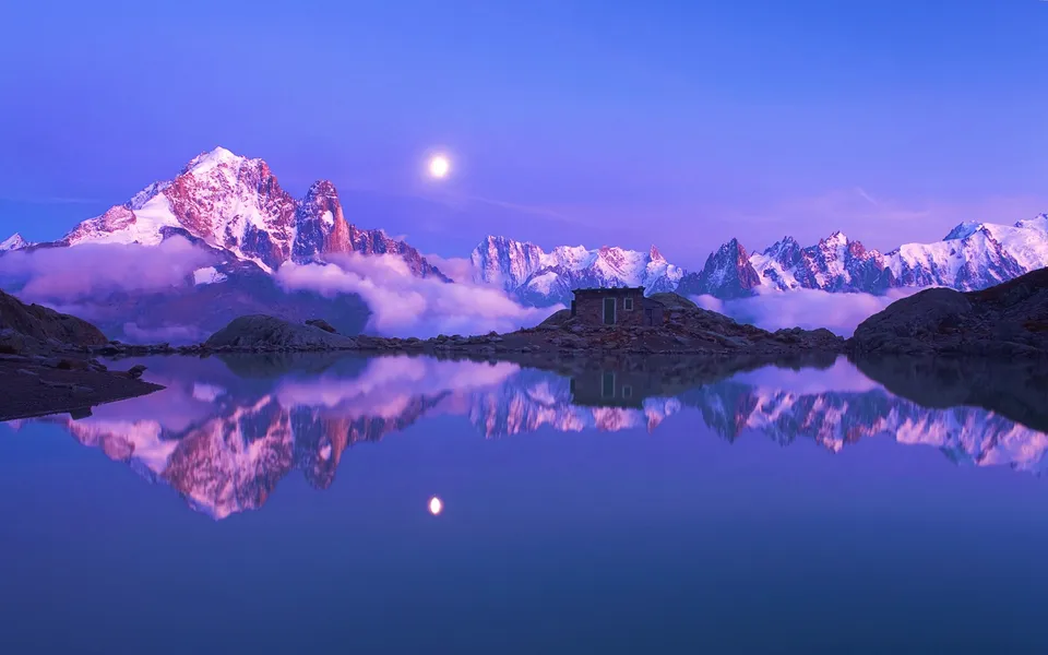 A serene alpine lake perfectly reflects snow-capped mountains and the moon under a twilight sky.