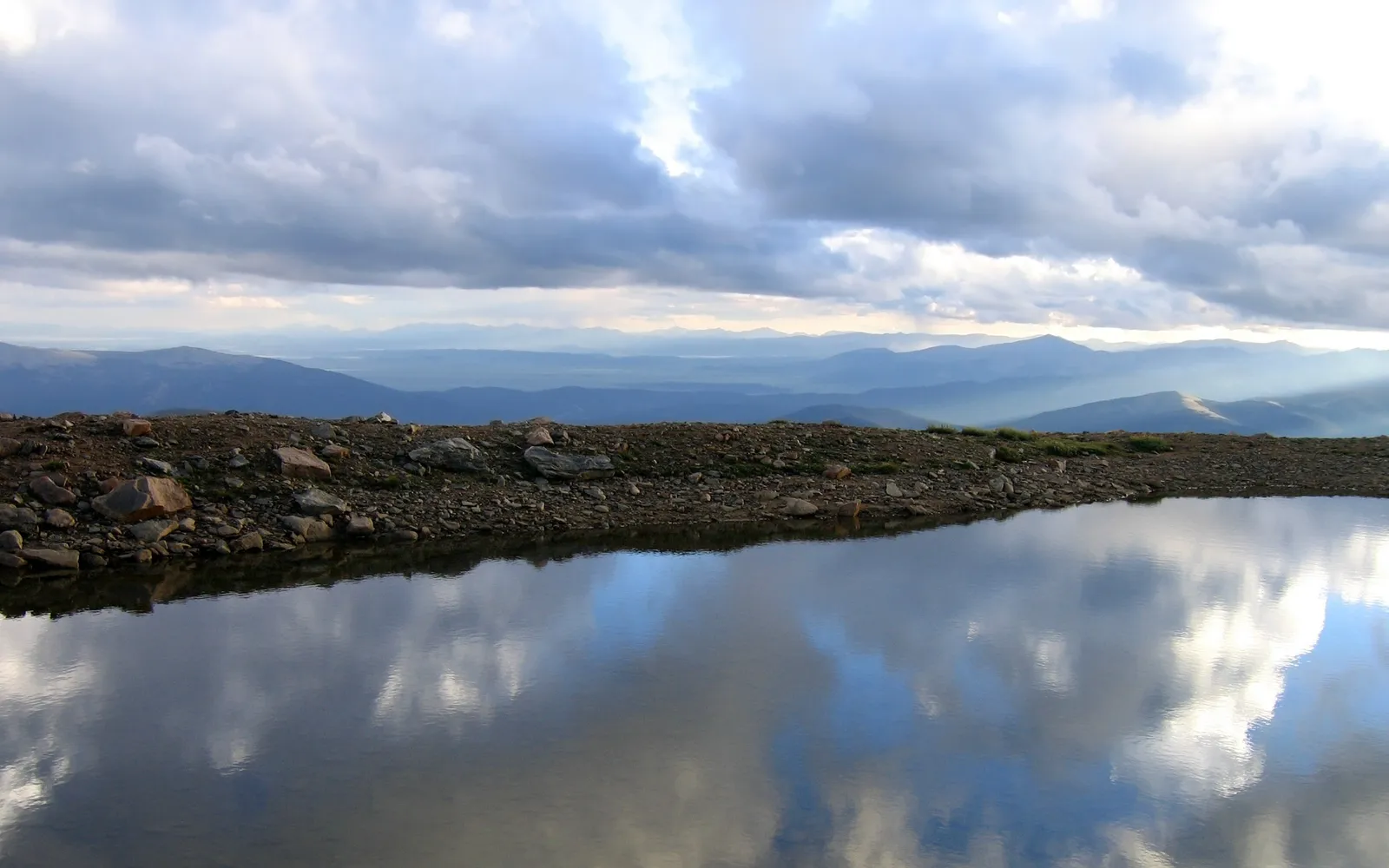 An alpine lake mirrors a cloudy sky with sunbeams breaking through, showing a rocky shore and distant mountains.