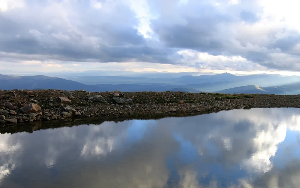 An alpine lake mirrors a cloudy sky with sunbeams breaking through, showing a rocky shore and distant mountains.