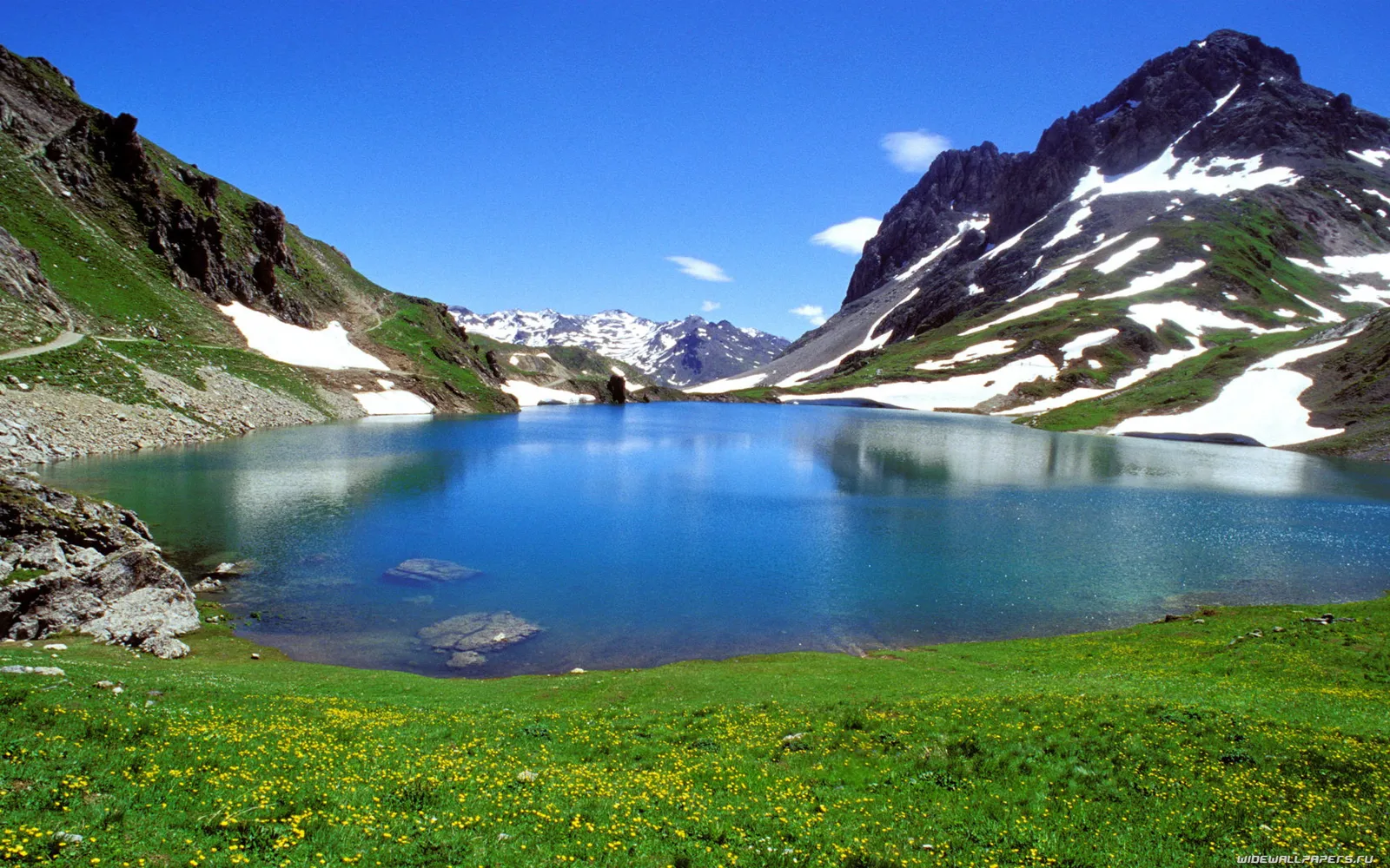 A bright blue alpine lake reflects the surrounding mountains, with patches of snow and green grass.
