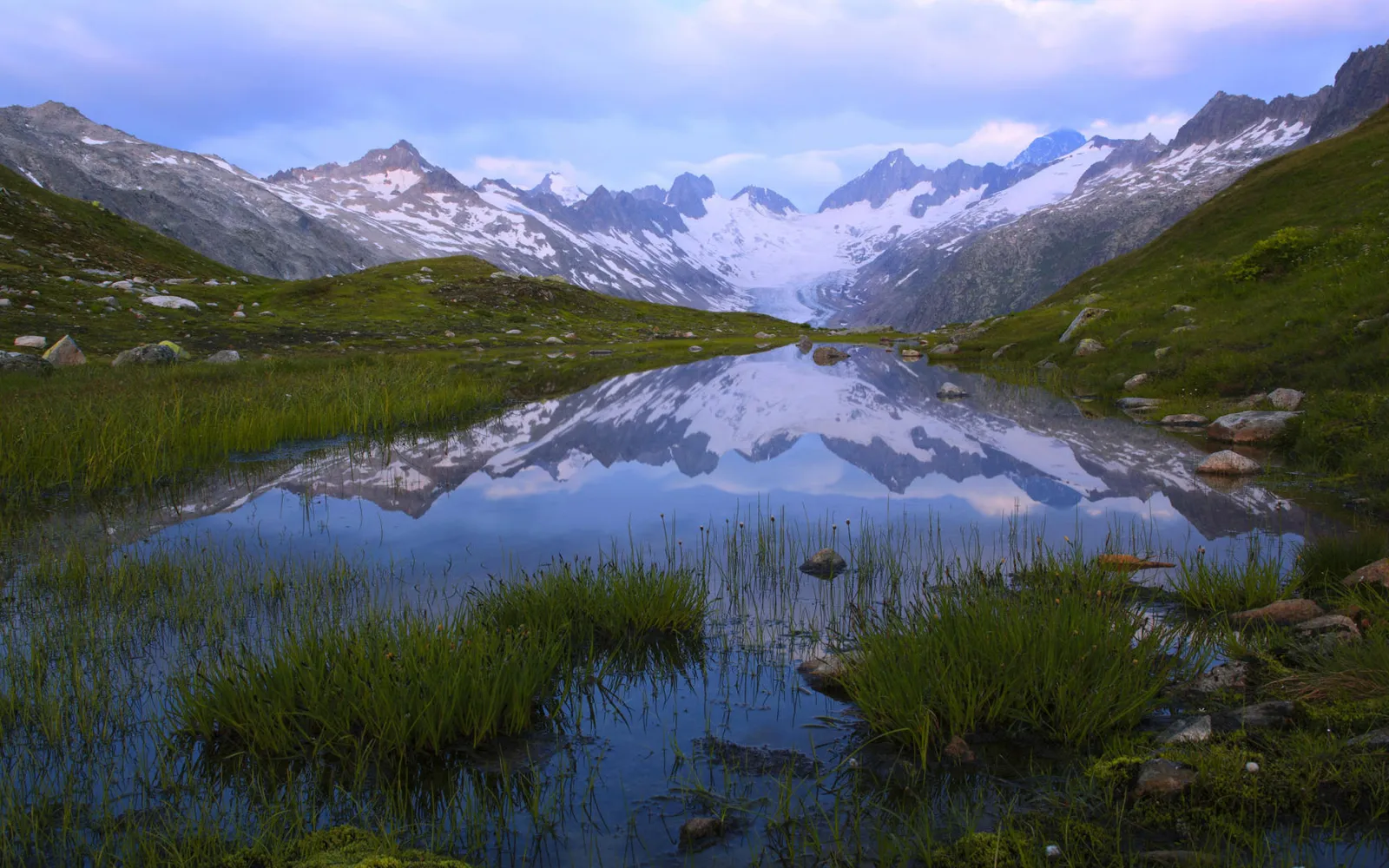 A calm alpine lake reflects snow-capped mountains under a cloudy sky, with green grass in the foreground.