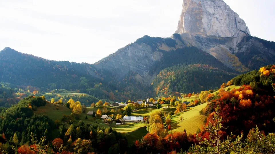 A picturesque village nestled in a valley with mountains in the background, surrounded by trees displaying autumn colors.