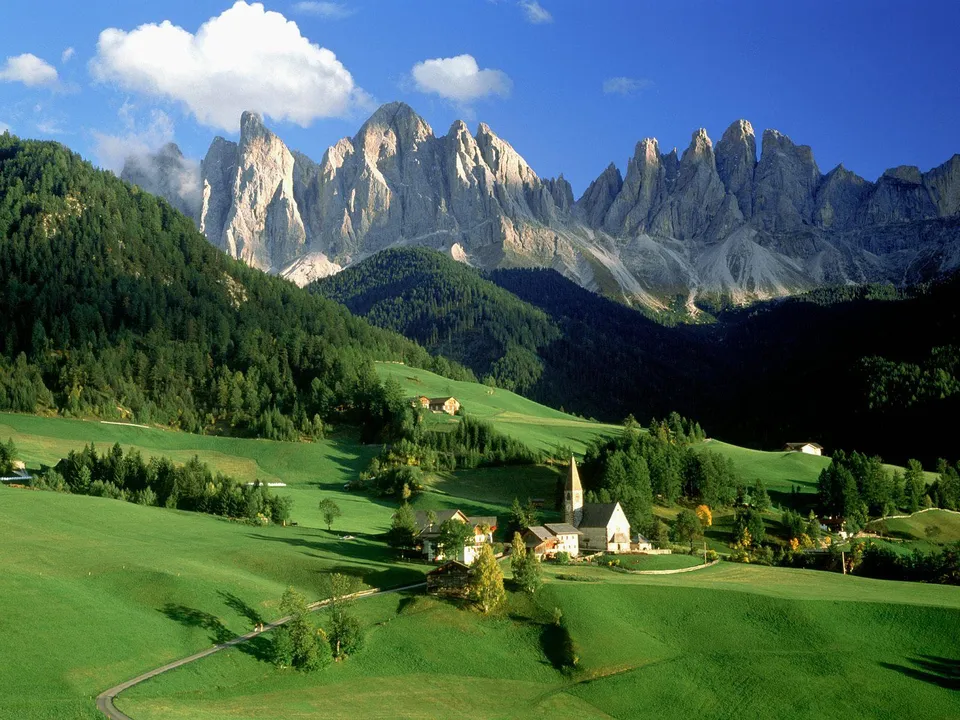 A picturesque village with a church sits in a lush green valley below dramatic, jagged mountain peaks under a blue sky with clouds.