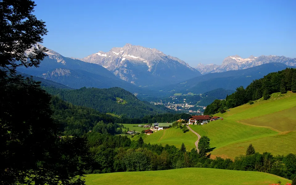 A picturesque village sits in a valley with green hills and snow-capped mountains in the background under a clear blue sky.