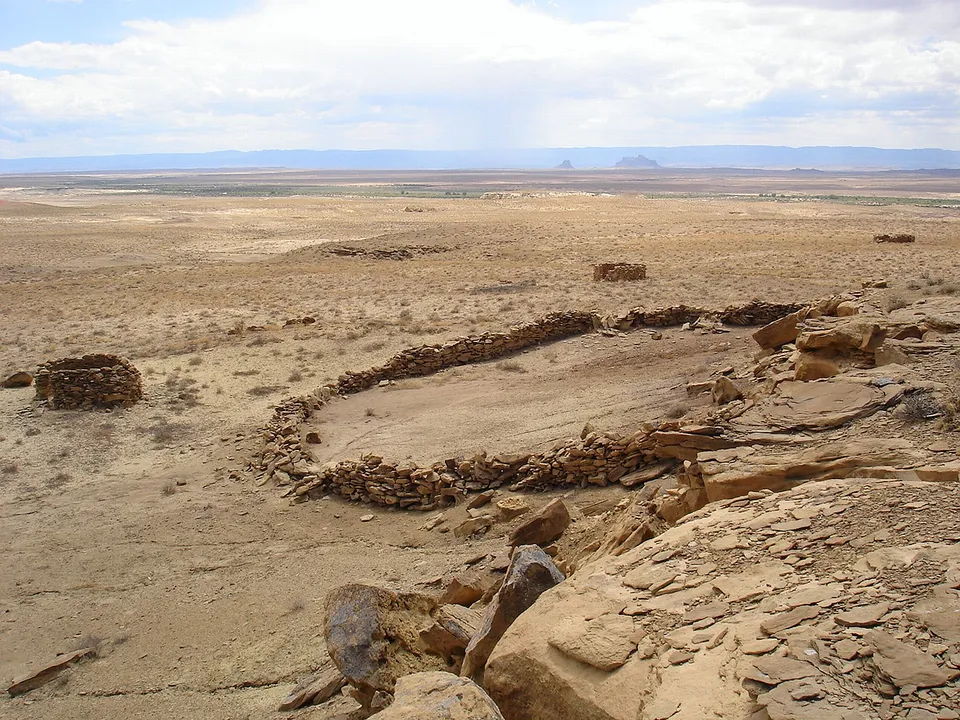 Ancient stone ruins are visible in a vast, dry desert landscape under a cloudy sky.