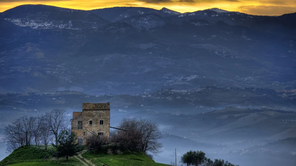 An old stone farmhouse sits on a grassy hill with bare trees, overlooking misty blue mountains under a golden sky.