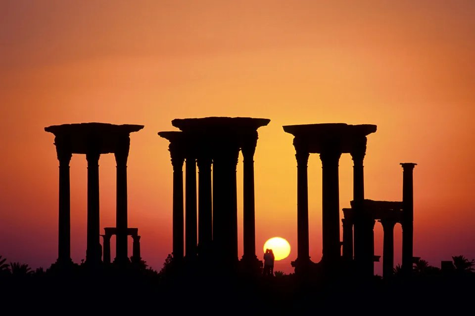 Silhouetted ancient Roman columns against a vibrant orange and yellow sunset sky.