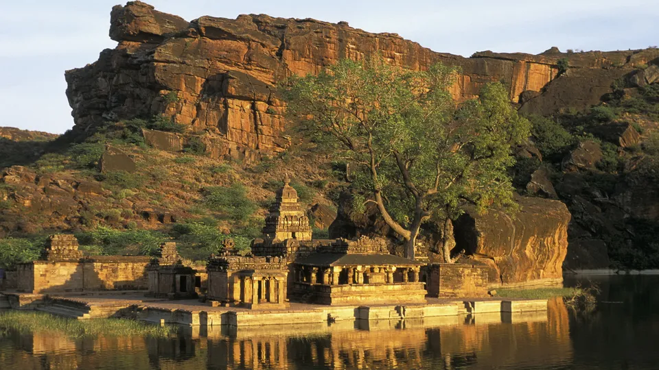 An ancient stone temple complex sits beside a calm body of water, with a large rock formation behind it.