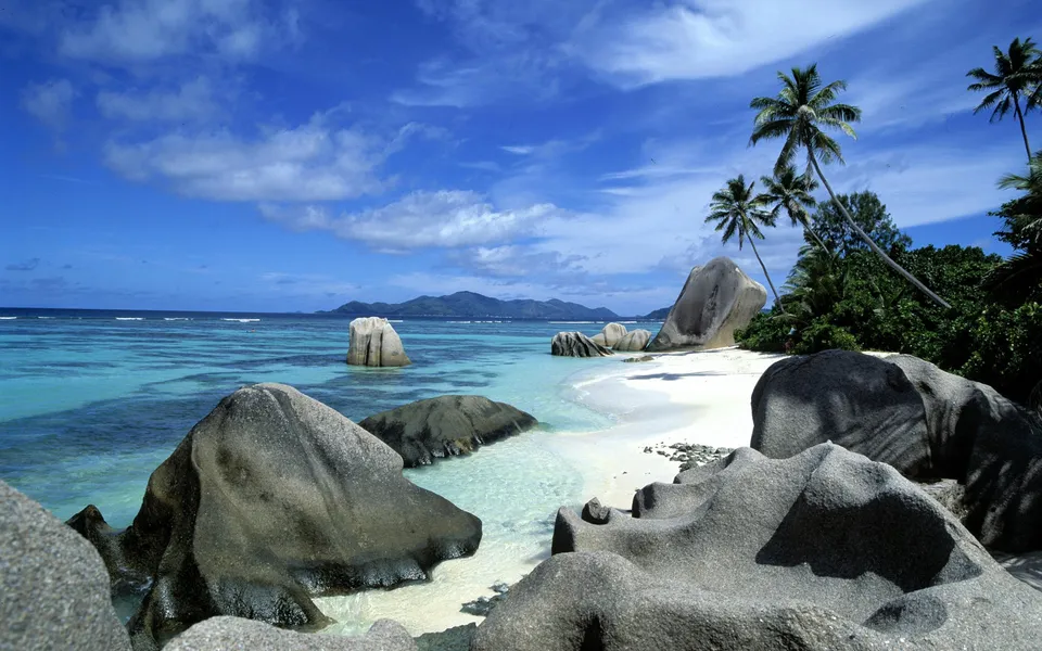 Large granite boulders line the white sand beach of Anse Source D'Argent, with turquoise water and palm trees.
