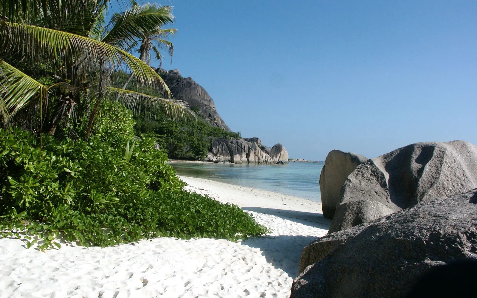 A view of a tropical beach with white sand, lush green vegetation, granite boulders, and clear turquoise water under a bright blue sky.