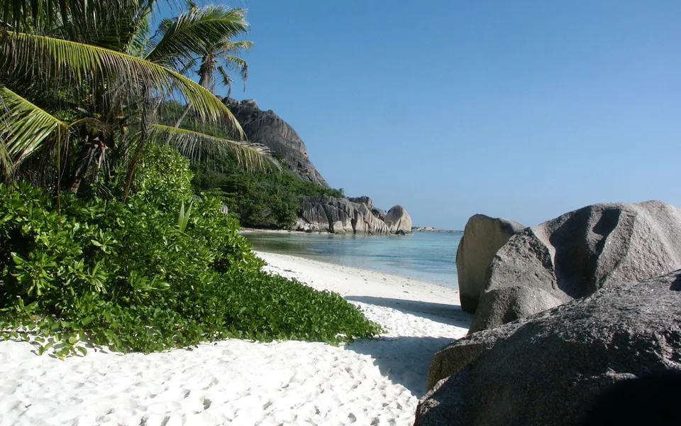 A view of a tropical beach with white sand, lush green vegetation, granite boulders, and clear turquoise water under a bright blue sky.