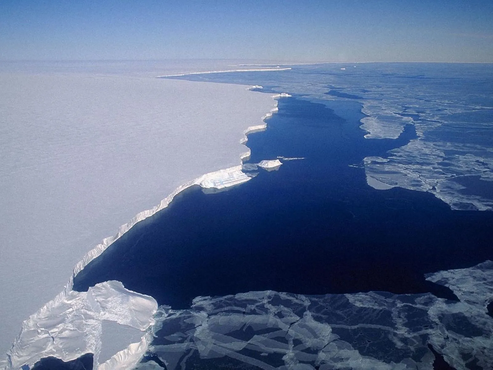 Aerial view of a vast white ice shelf meeting the dark blue ocean with scattered ice floes.