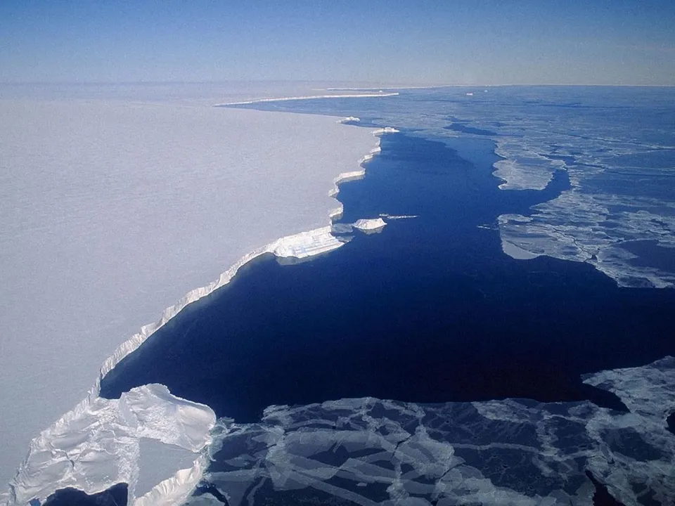 Aerial view of a vast white ice shelf meeting the dark blue ocean with scattered ice floes.