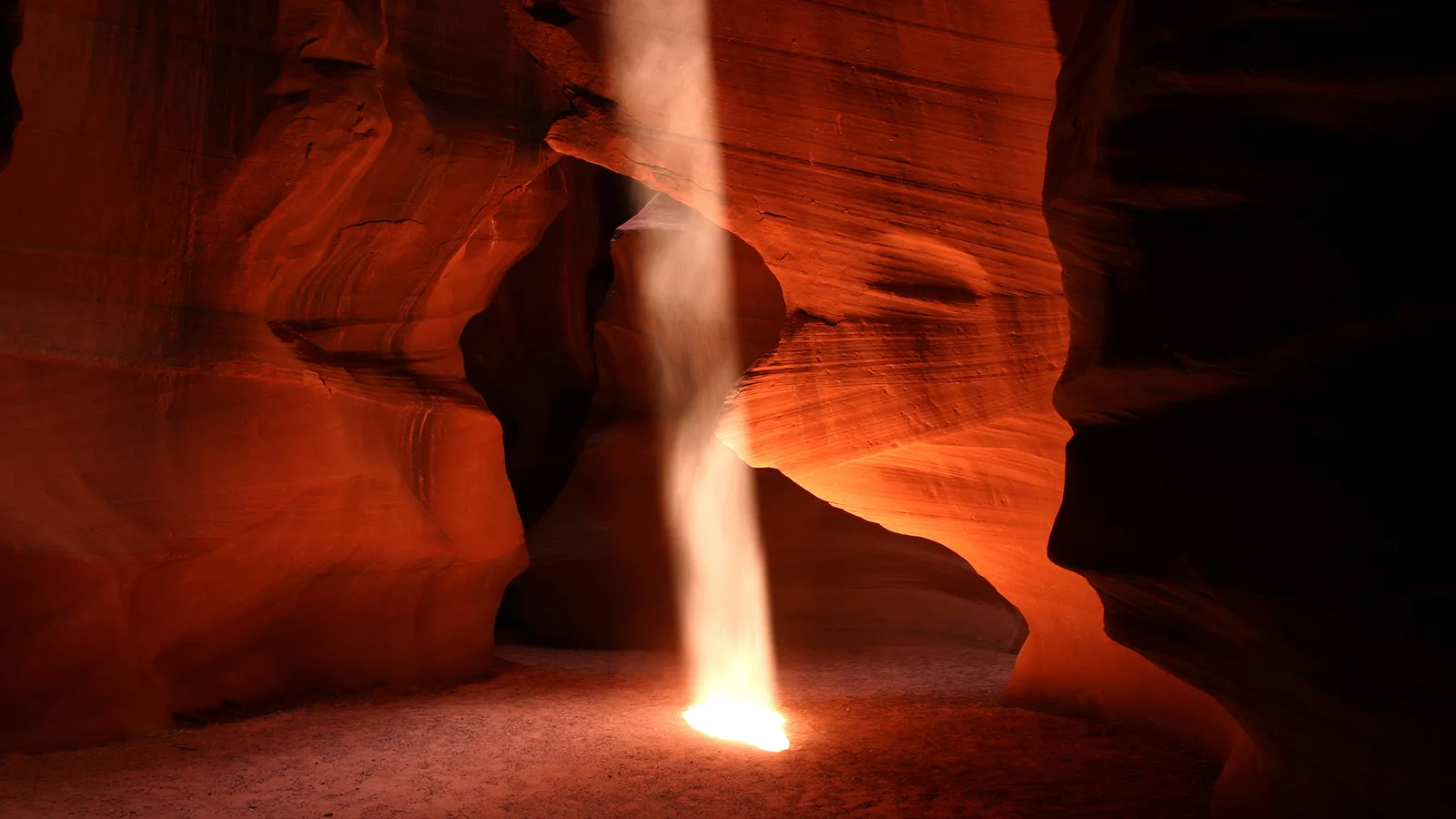 A dramatic beam of light shines down from above into a narrow slot canyon, illuminating the sandy floor.