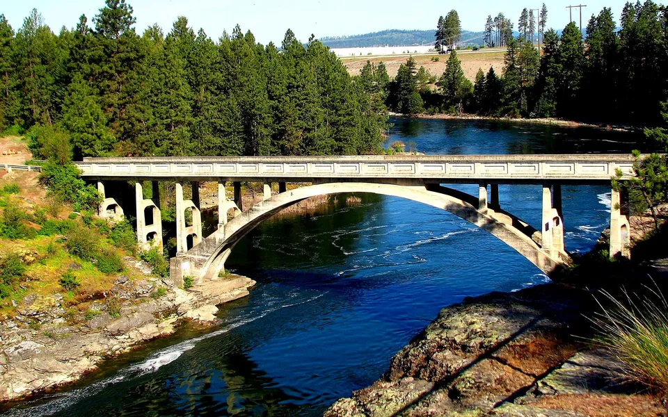 A concrete arch bridge crosses a wide, blue river surrounded by green trees and rocky shores.