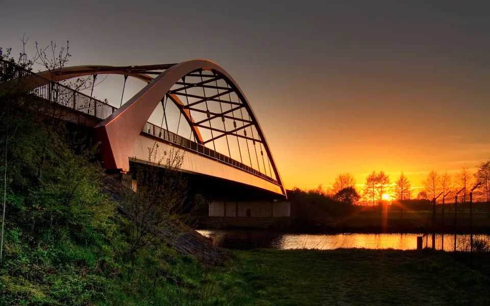 An arch bridge spans over a calm river during a vibrant sunset with trees silhouetted against the orange sky.