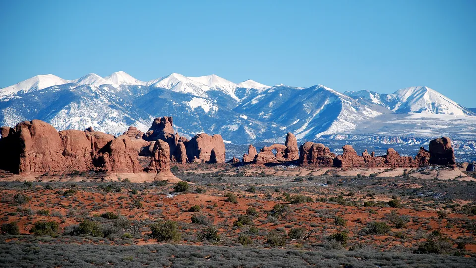 A vast desert landscape with red rock formations in the foreground and snow-covered mountains in the background under a clear blue sky.