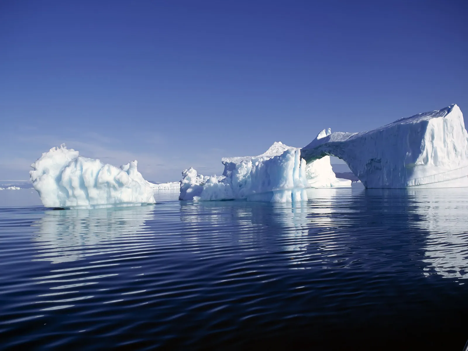 A large iceberg with a natural archway formation floats in the calm Arctic ocean under a bright blue sky.