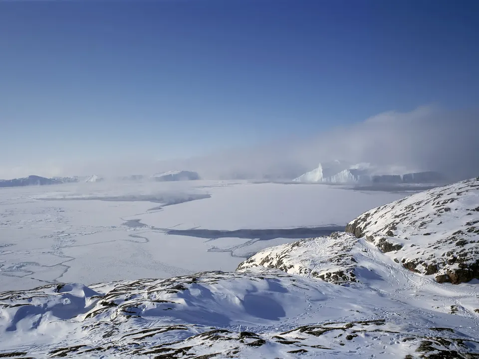 A vast frozen sea with icebergs and mist under a clear blue sky, viewed from a snow-covered rocky outcrop.