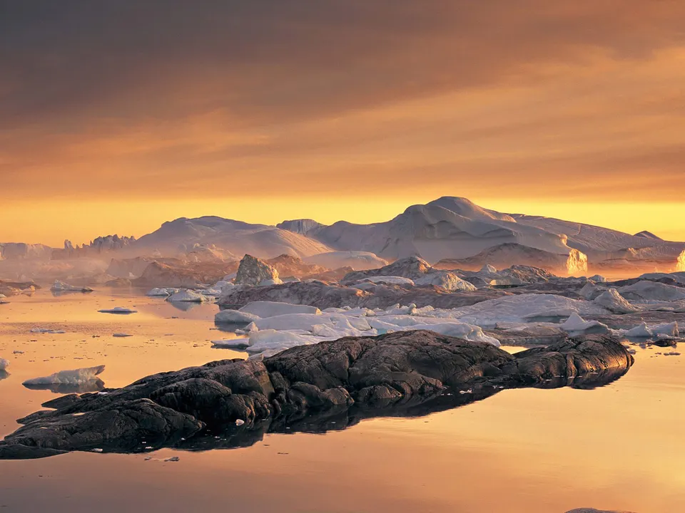 Icebergs float in calm water with snow-capped mountains in the background under a golden sky.