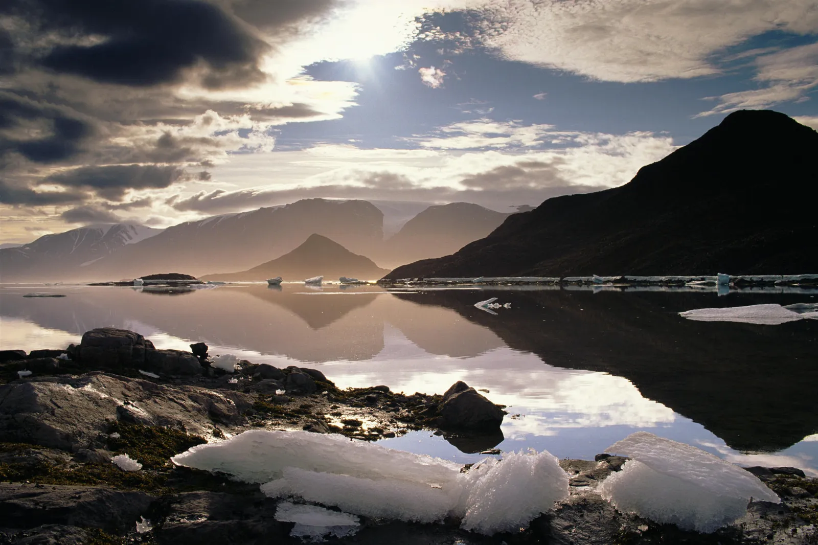 A calm lake with ice chunks in the foreground reflects dark mountains under a cloudy sky with sunbeams.