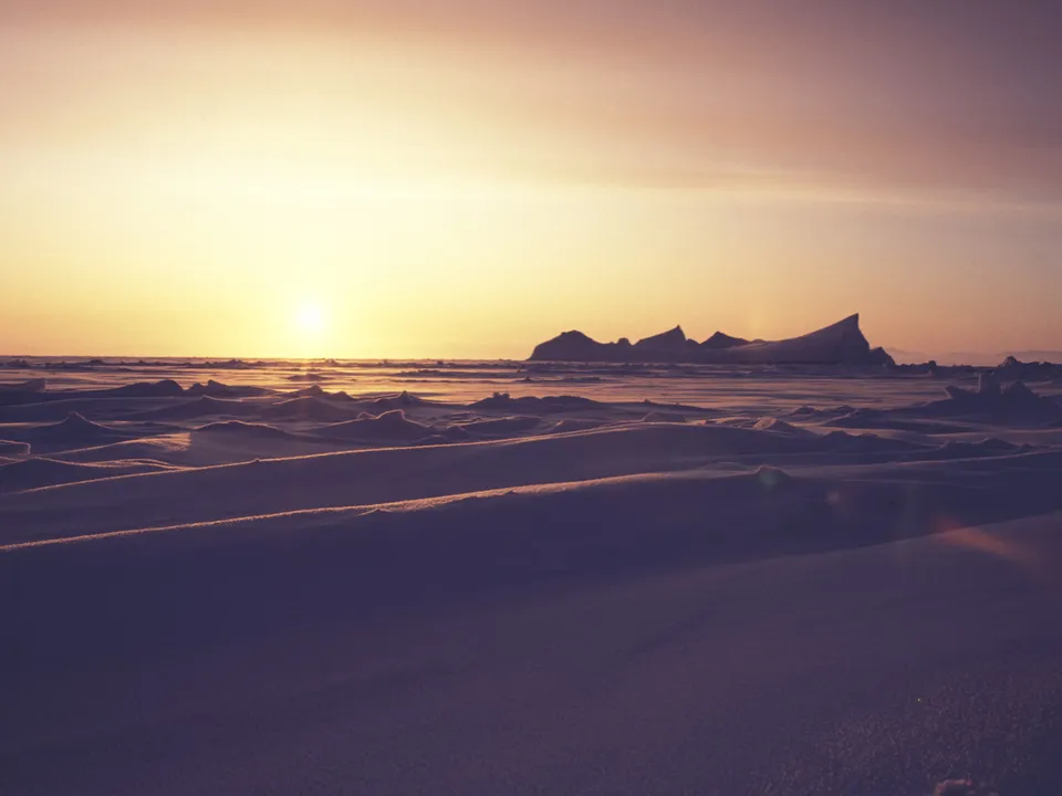 A low arctic sun casts a warm glow across a frozen, undulating landscape of snow and ice.