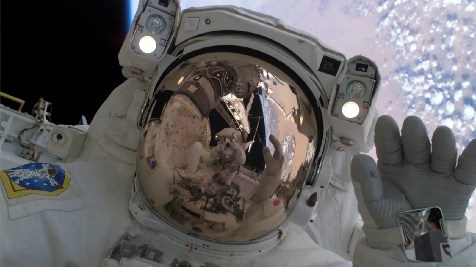 An astronaut in a white spacesuit takes a selfie with the Earth and clouds visible in the background.
