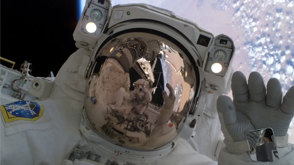 An astronaut in a white spacesuit takes a selfie with the Earth and clouds visible in the background.