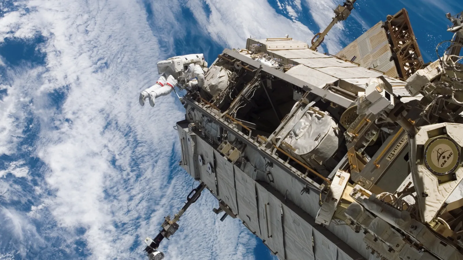 An astronaut in a white spacesuit is outside the International Space Station, with Earth and clouds visible below.