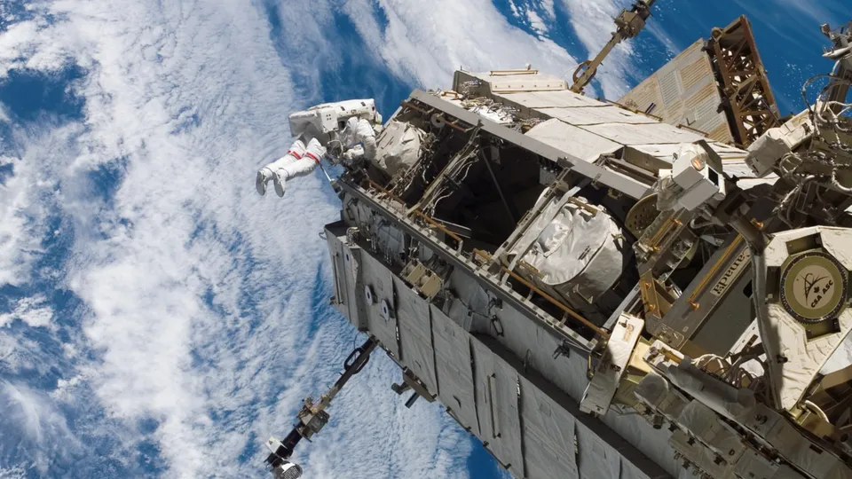 An astronaut in a white spacesuit is outside the International Space Station, with Earth and clouds visible below.